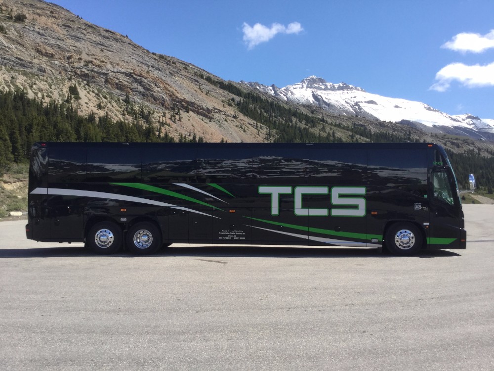 Black TCS Canada charter bus in front of snowcapped mountains on sunny mountain highway